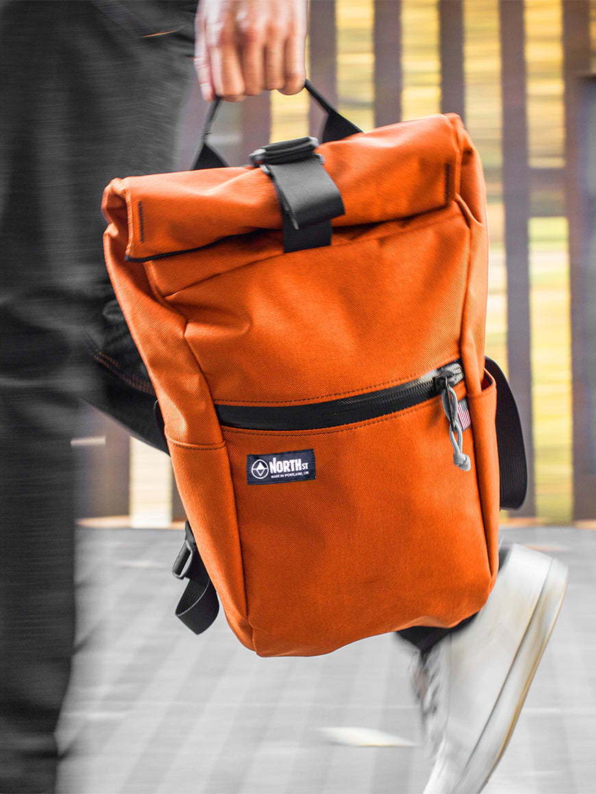 Orange backpack with a visible brand logo held by a person on a blurred background all-groups