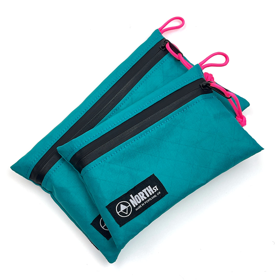 Teal travel pouches with black zippers and pink drawstrings on a white background