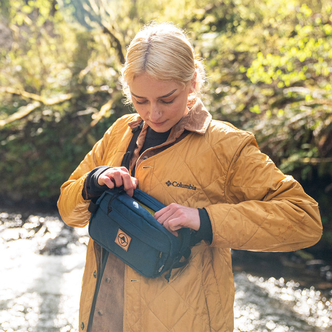woman on a forest patch reaching into a blue sling pack