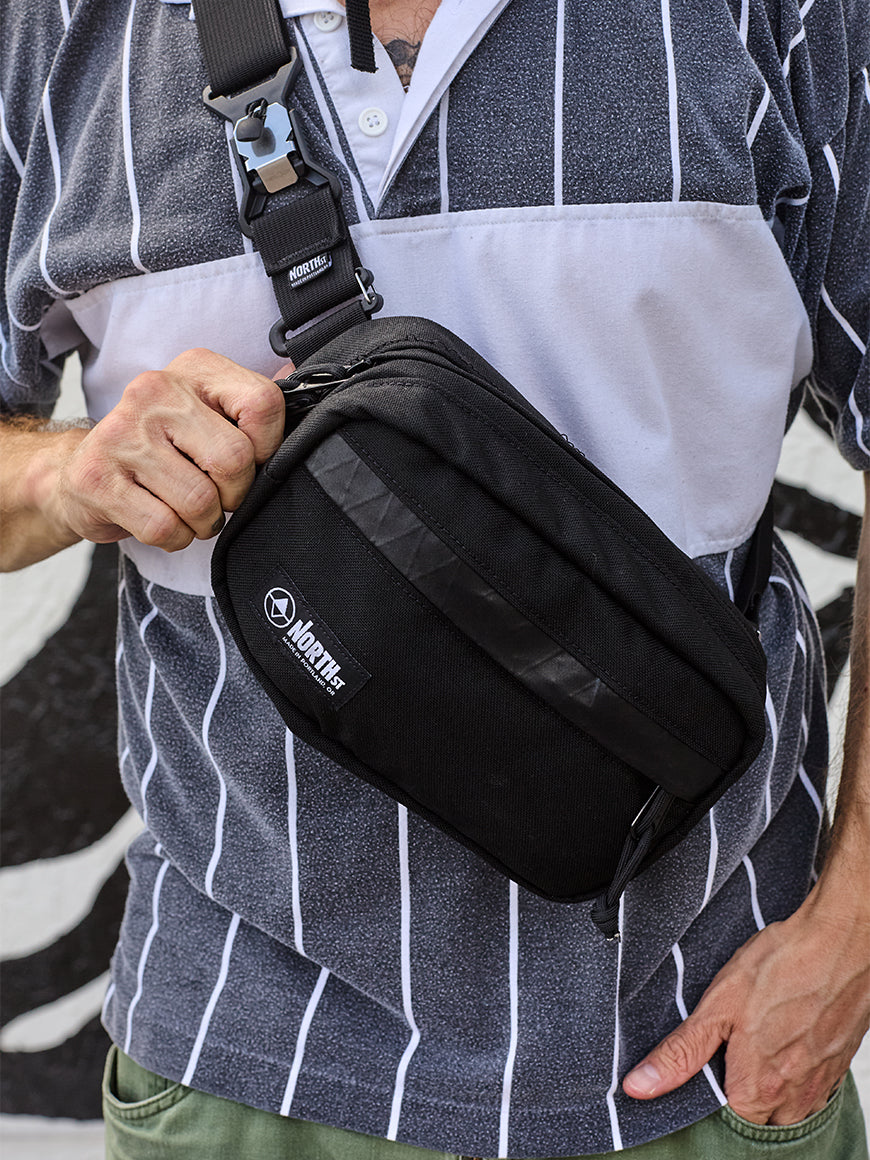 Man wearing a black waist bag with a visible brand logo, standing outdoors.